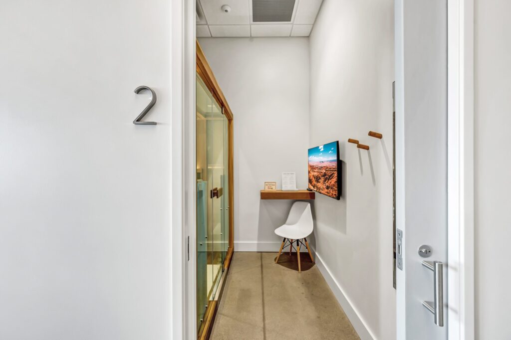 Long, narrow treatment room at Perspire Sauna Studio in Peachtree Corners. The floor is light brown, walls are white. There's a white chair and small wall shelf at the end of the room, across from a sauna. The number 2 is on the wall outside the rooms open door.