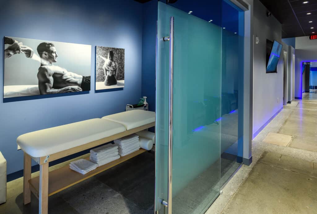 A treatment room and hallway at Icebox Cryotherapy studio in Peachtree Corners. There's a massage-style table in the center of the room and sliding glass doors to close off the room. The walls are blue and there are black and white photo prints for decoration. Rolled up white towels are on a shelf below the table.