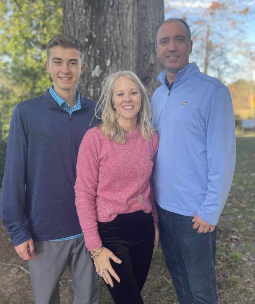 A blonde haired woman in a pink, long sleeve shirt standing next to her teenage son and husband.