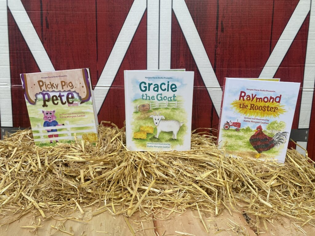 Three children's books: one about a pig, one about a rooster and one about a goat, sitting on top of a bale of hay in front of a barn.