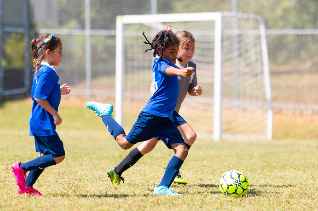 Three kids in blue jerseys on a soccer field playing a match. The kid in front has their leg pulled back, getting ready to kick the ball.