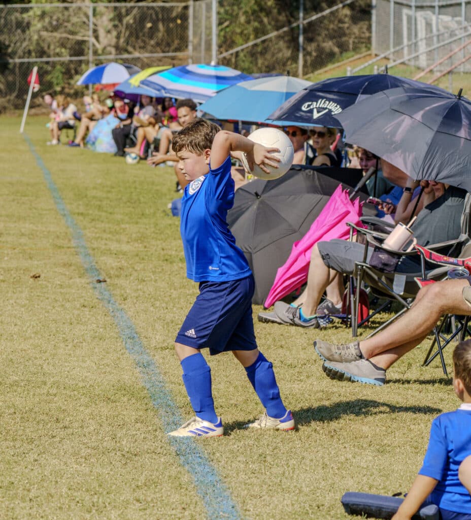 Families sitting in outdoor chairs under umbrellas along the side of a soccer field, watching their kids play.