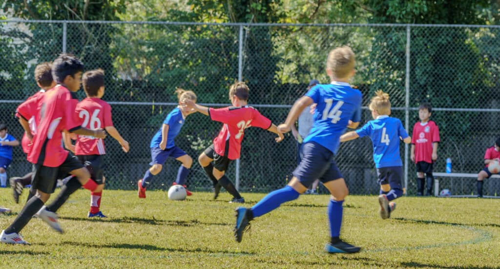 Kids playing in a soccer match, some in red jerseys and some in blue. They're running on the field and one has the ball.