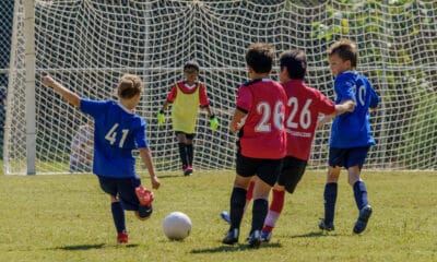 A group of kids, some in blue jerseys and some in red, playing soccer. Number 41 is kicking the ball towards the goal, where the other team's goalie waits.