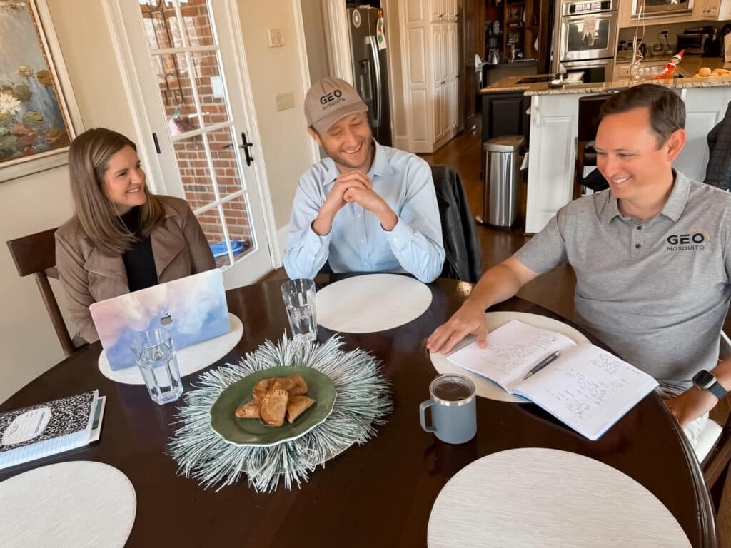 A woman and two men sitting at a kitchen table with snacks, drinks and notebooks discussing plans for business expansion.