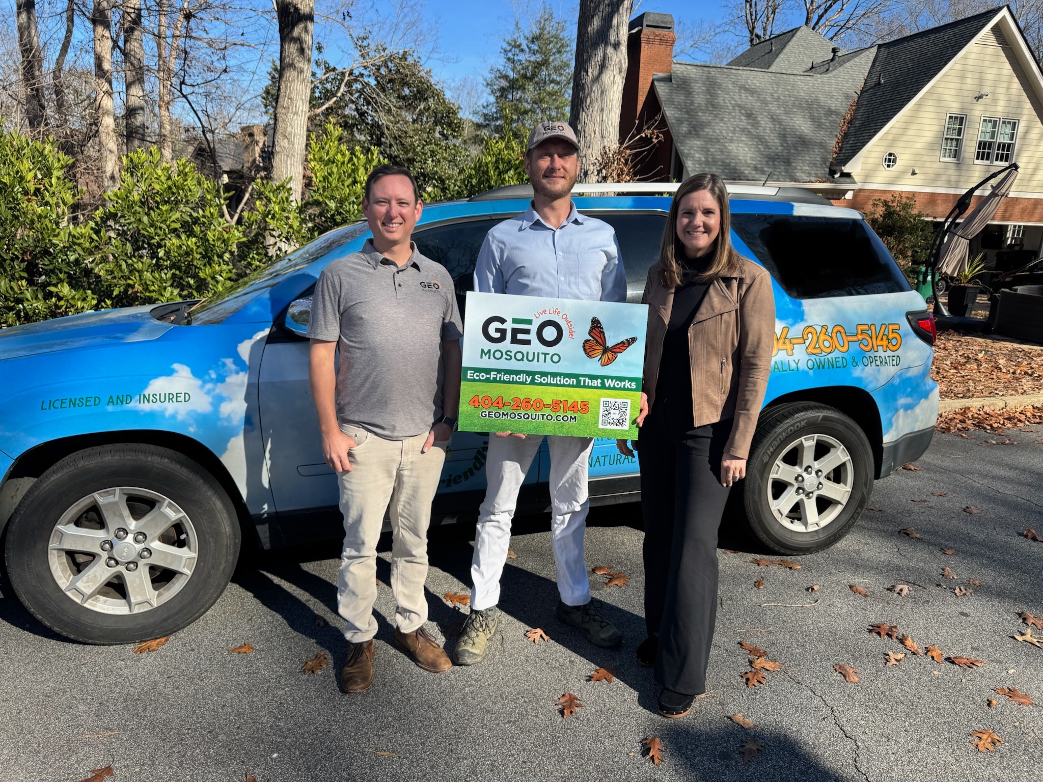 Two men and one woman standing next to a blue SUV with company logo on it and holding a sign for Geo Mosquito.