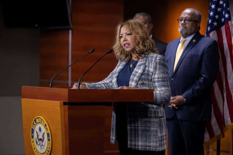 Rep. McBath, of Georgia's 6th District, at a podium speaking to an audience. A man and woman stand behind her, along with an American flag.