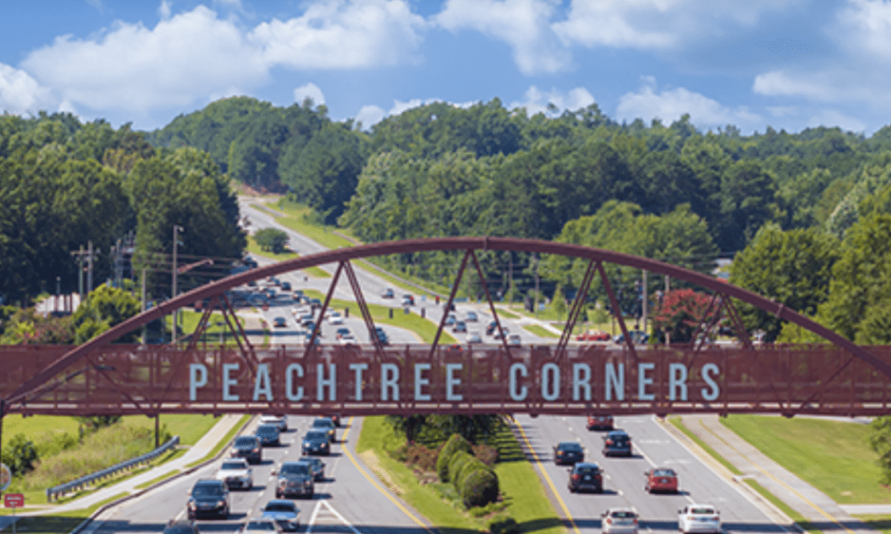 Aerial photo of the Peachtree Corners pedestrian bridge with trees in the background, businesses alongside, and cars driving on the six-lane road beneath it.