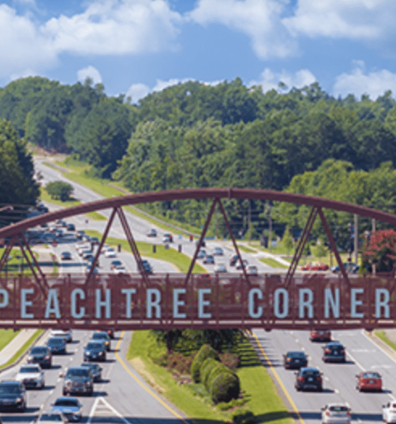 Aerial photo of the Peachtree Corners pedestrian bridge with trees in the background, businesses alongside, and cars driving on the six-lane road beneath it.