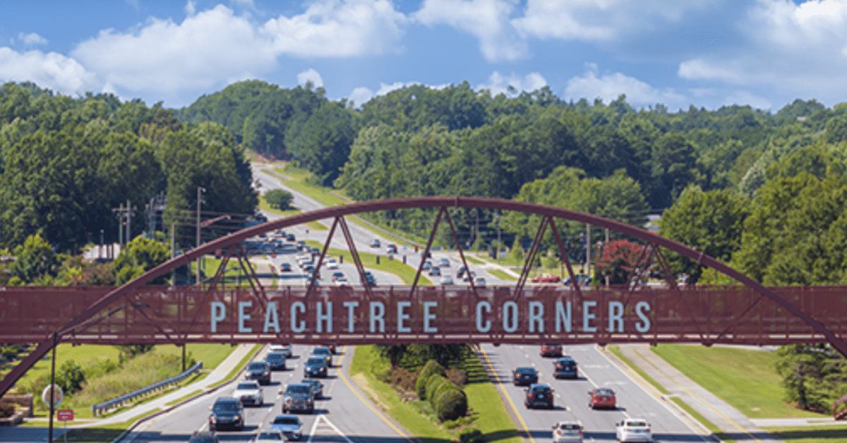 Aerial photo of the Peachtree Corners pedestrian bridge with trees in the background, businesses alongside, and cars driving on the six-lane road beneath it.