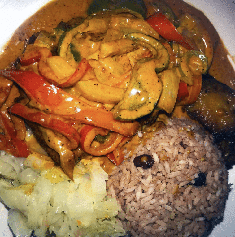 A plate of Jamaican food with black beans and rice, steamed green vegetable and a thick curry with meat and peppers