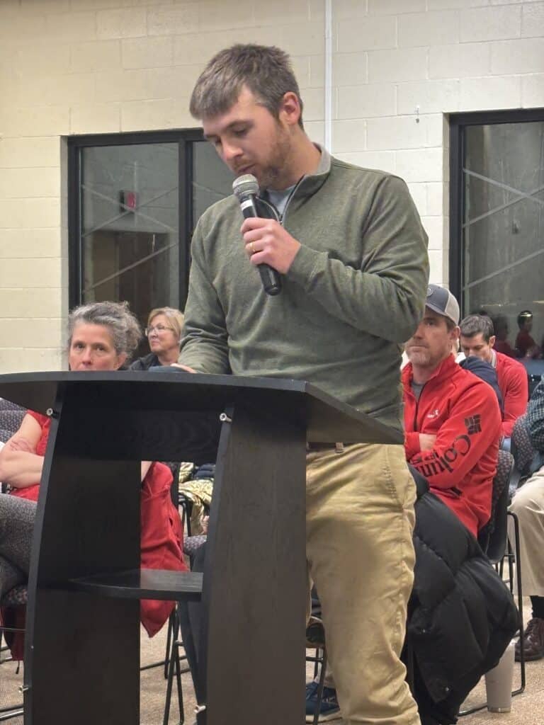 A man in an olive green long-sleeved shirt and khaki pants speaks from the audience podium at a community meeting