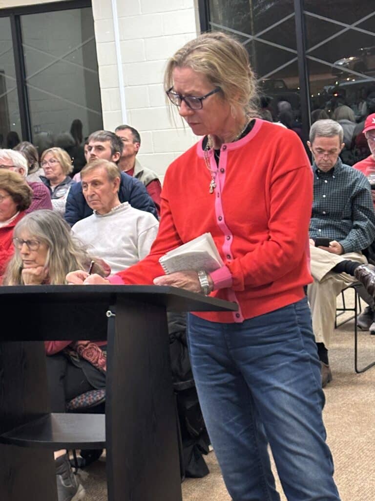 Middle-aged woman wearing a red sweater speaking from an audience podium at a community meeting