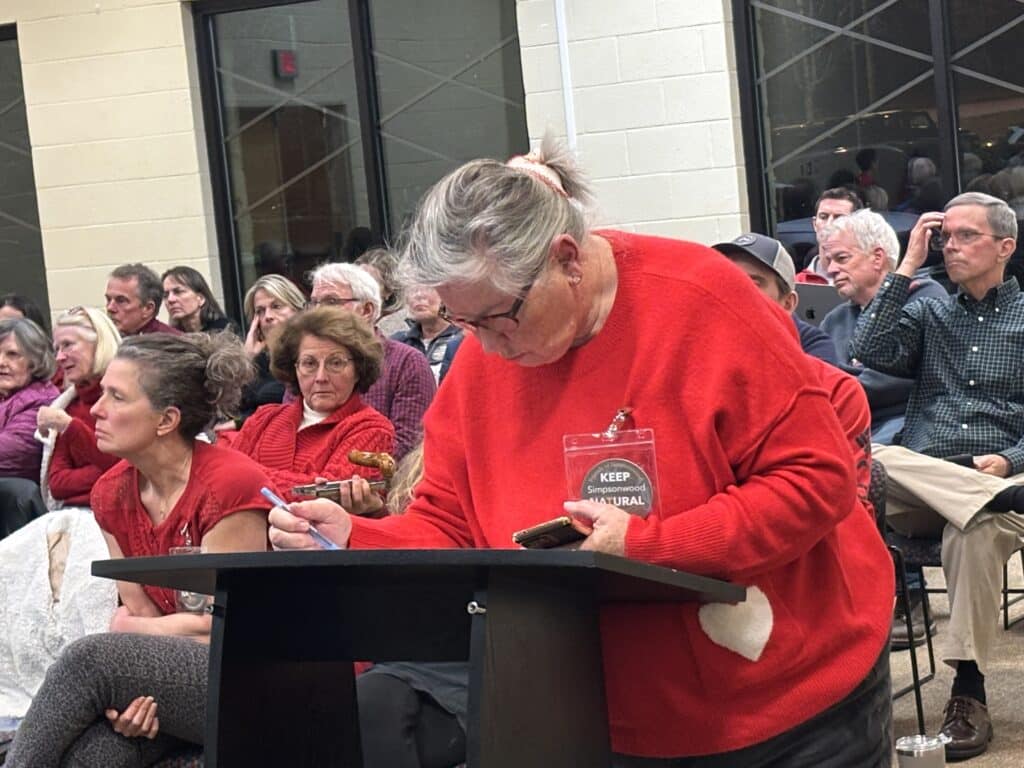 A grey-haired woman in a red sweater at a community meeting, signing up to speak at the podium