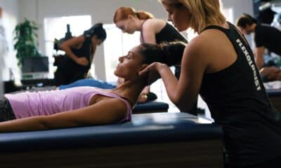 A woman with dark hair and pink top is lying on a massage-type table with her head lifted. Another woman, wearing a fitness top, is cradling her head to help stretch her neck.