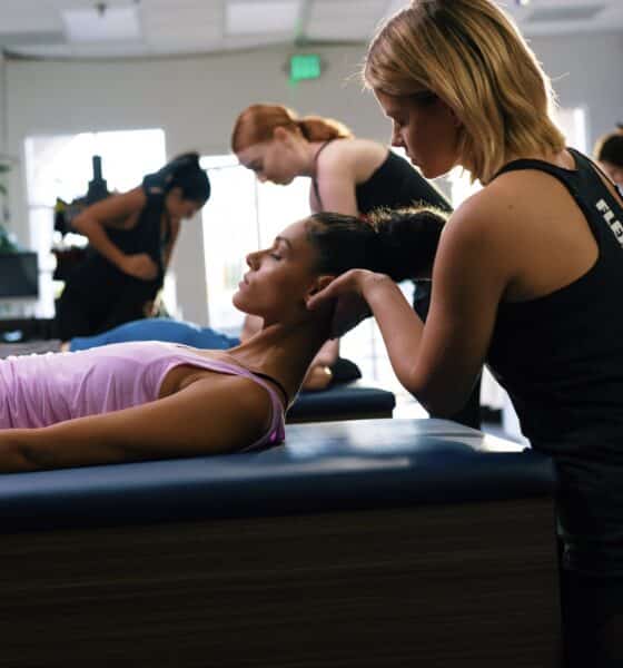 A woman with dark hair and pink top is lying on a massage-type table with her head lifted. Another woman, wearing a fitness top, is cradling her head to help stretch her neck.