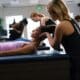 A woman with dark hair and pink top is lying on a massage-type table with her head lifted. Another woman, wearing a fitness top, is cradling her head to help stretch her neck.