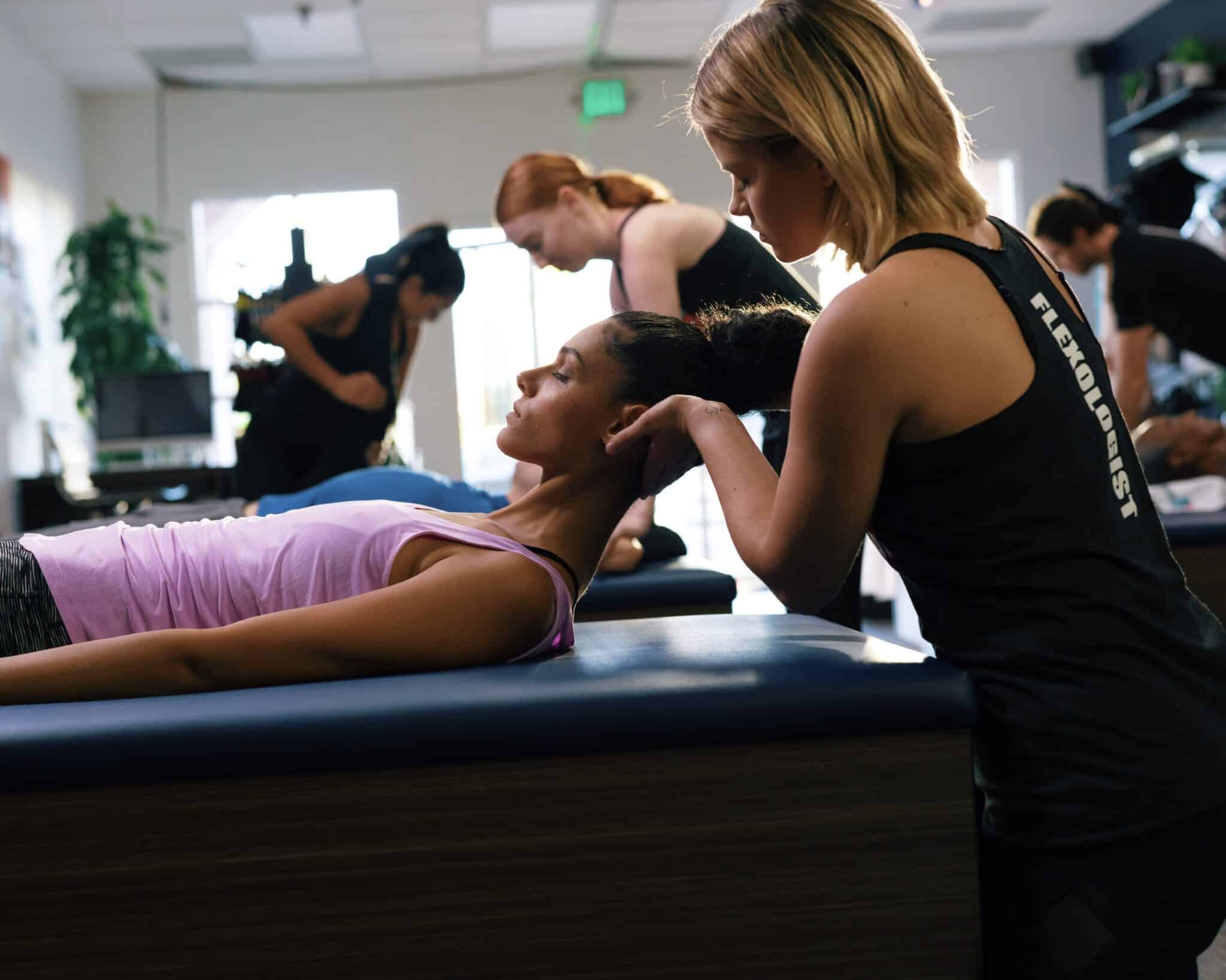 A woman with dark hair and pink top is lying on a massage-type table with her head lifted. Another woman, wearing a fitness top, is cradling her head to help stretch her neck.