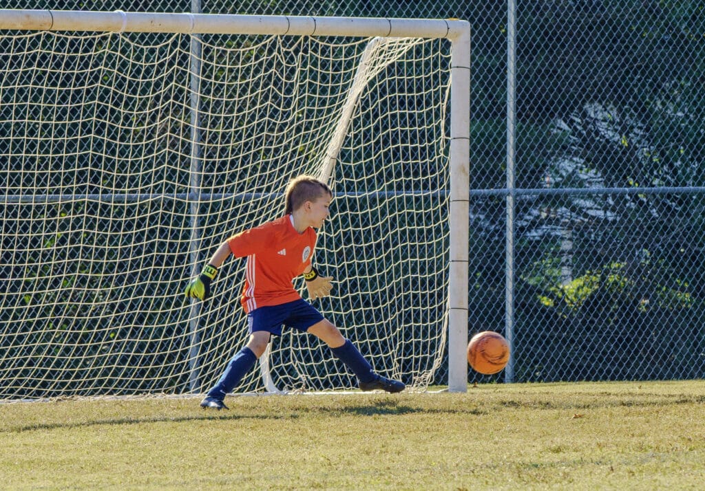 Goalie on a youth soccer team, wearing a red jersey, blocking a soccer ball with his foot