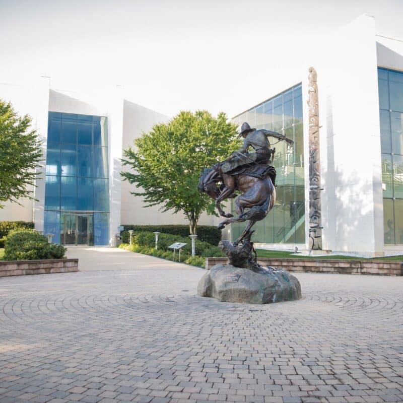 Modern building with a few green trees, shrubs, brick courtyard and a cowboy statue.