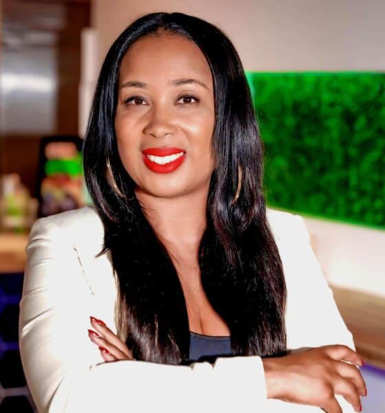 A young Black woman with long straight hair and red lipstick, wearing a white suit and standing with her arms crossed in front of her as she smiles at the camera.