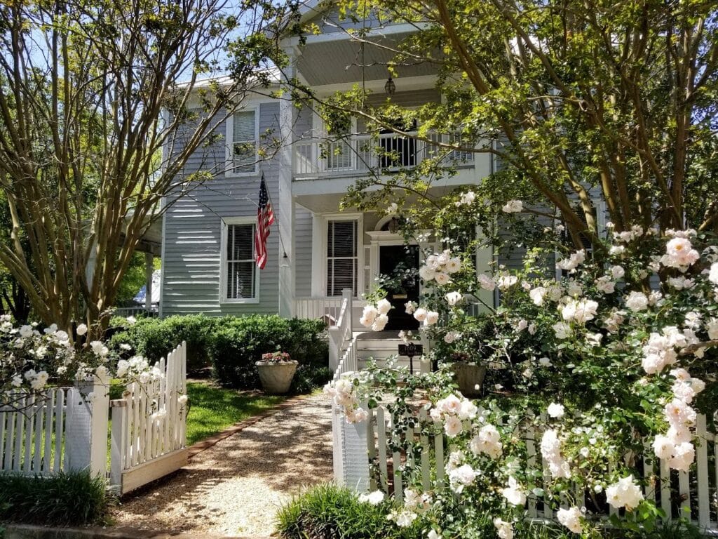 Historic two-story wooden home in Madison, GA surrounded by trees, a lush flower garden and a white picket fence.