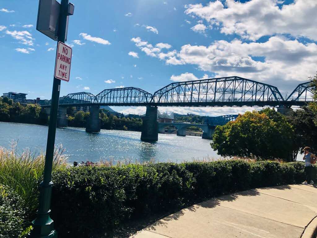 Large bridge in the distance crossing the river in Chattanooga, TN. The water is clear, the sky is blue with white clouds and there's a sidewalk, bushes and sign in the foreground.