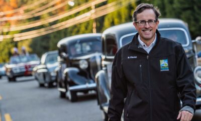 A white man with glasses, wearing a dark colored jacket walking in the road alongside a line of vintage cars.