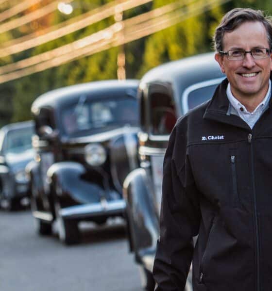 A white man with glasses, wearing a dark colored jacket walking in the road alongside a line of vintage cars.