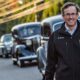 A white man with glasses, wearing a dark colored jacket walking in the road alongside a line of vintage cars.