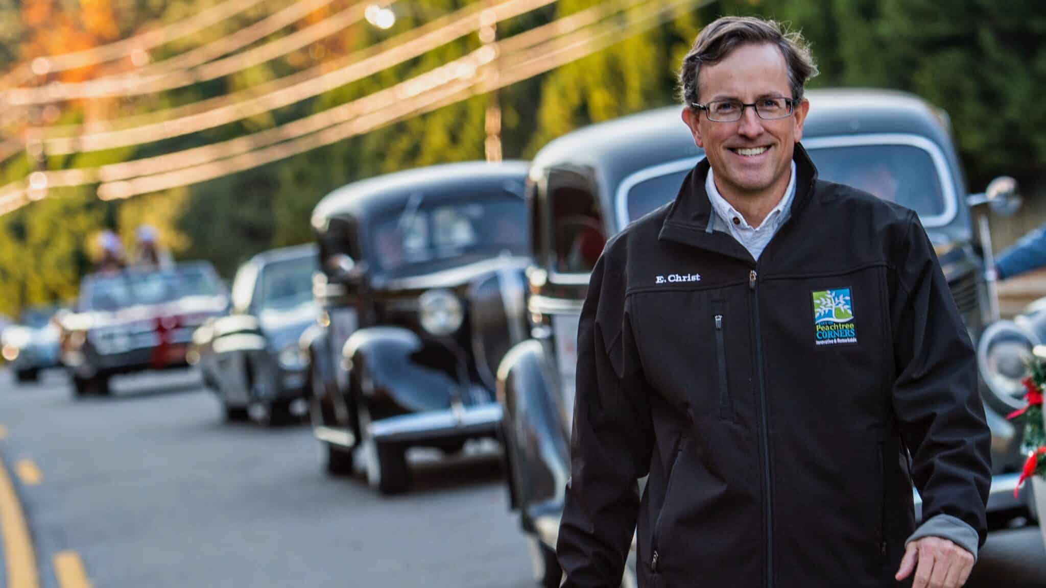 A white man with glasses, wearing a dark colored jacket walking in the road alongside a line of vintage cars.