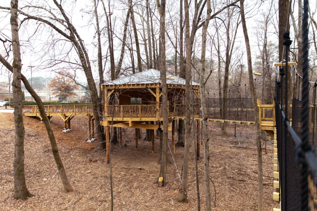 Wooden treehouse and walkway, just elevated off the ground, in a wooded area in fall. The treehouse is surrounded by leaf-less trees. Brown leaves cover the ground.