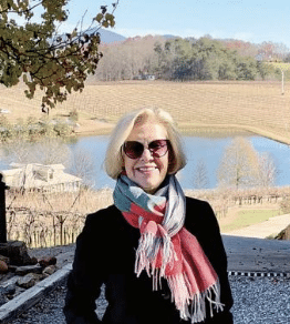 A smiling, middle-aged woman with short blonde hair, wearing sunglasses, a colorful scarf and black, long-sleeved top. She's standing outdoors in front of a small pond/lake in fall, with trees and hills in the background.