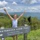 A young white girl wearing a blue sleeveless top standing next to a trail sign on top of a hiking path with her arms raised overhead. The trees behind her in the background are lush and green and the sky overhead is blue and dotted with fluffy white clouds.