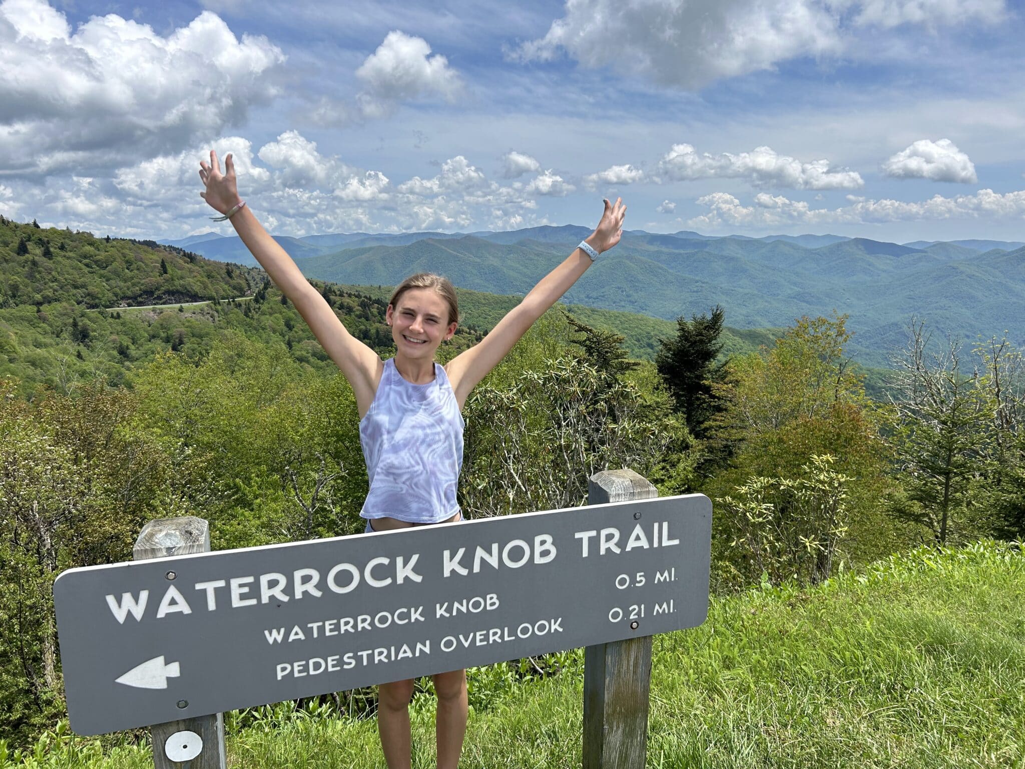 A young white girl wearing a blue sleeveless top standing next to a trail sign on top of a hiking path with her arms raised overhead. The trees behind her in the background are lush and green and the sky overhead is blue and dotted with fluffy white clouds.