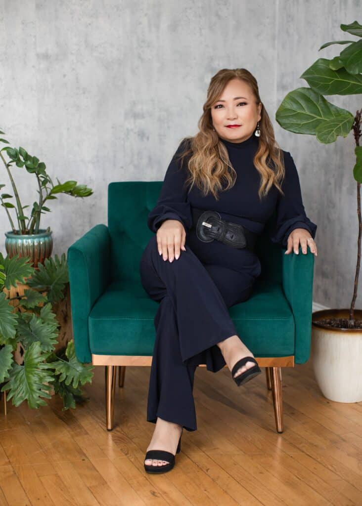 A middle-aged Korean woman with long hair, wearing black and sitting in a modern turquoise and gold chair next to potted plants