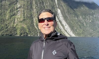 A man with short dark and grey hair wearing sunglasses and a black, long-sleeved zip up. He's standing by the railing of a boat in Milford Sound New Zealand with water and tall cliffs behind him. He's smiling and the sky above is dark blue with a few wispy white clouds.