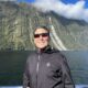 A man with short dark and grey hair wearing sunglasses and a black, long-sleeved zip up. He's standing by the railing of a boat in Milford Sound New Zealand with water and tall cliffs behind him. He's smiling and the sky above is dark blue with a few wispy white clouds.