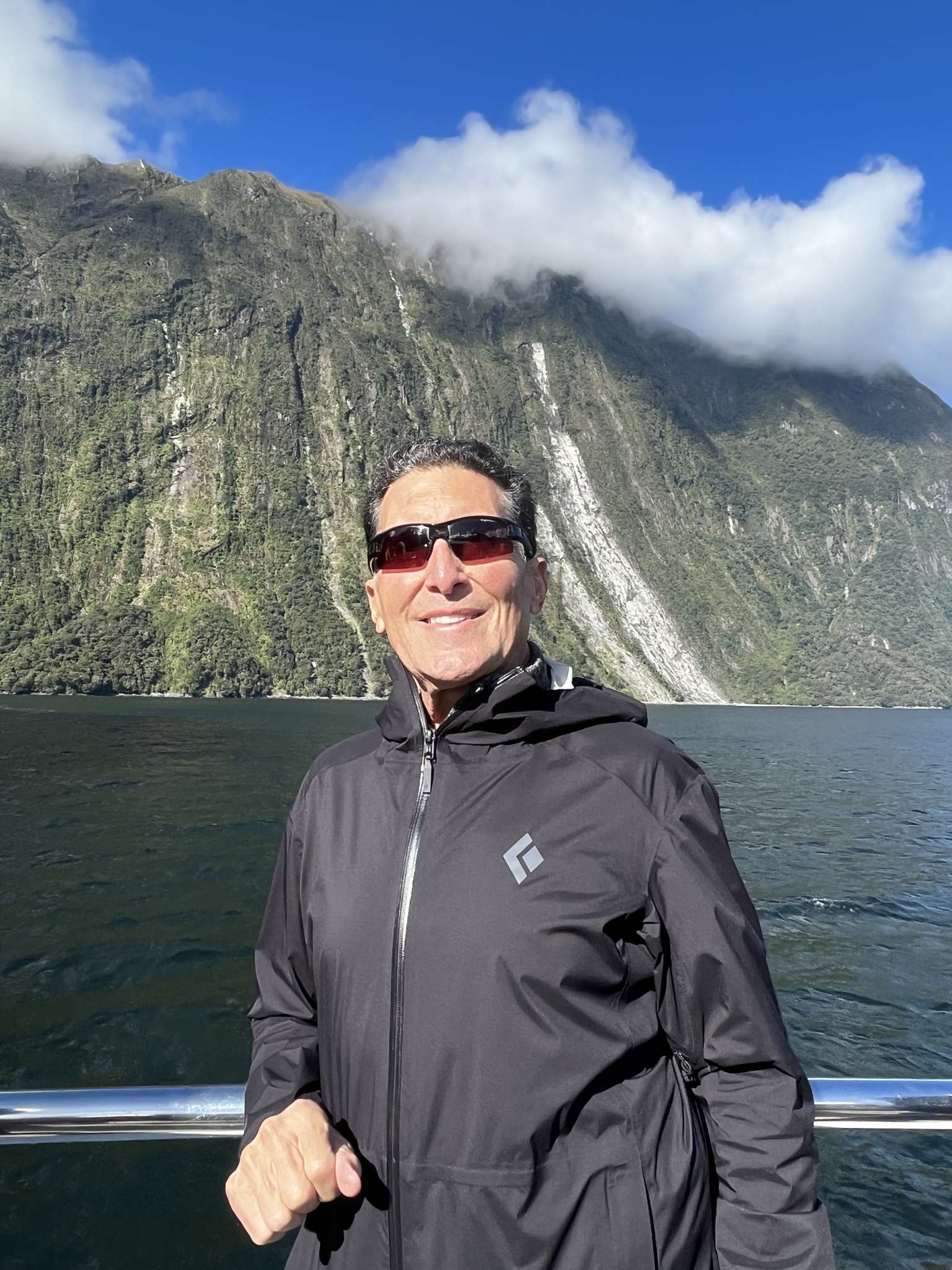 A man with short dark and grey hair wearing sunglasses and a black, long-sleeved zip up. He's standing by the railing of a boat in Milford Sound New Zealand with water and tall cliffs behind him. He's smiling and the sky above is dark blue with a few wispy white clouds.