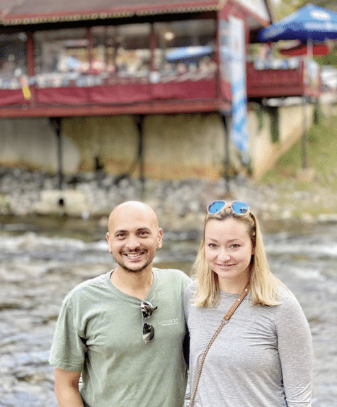 A man and woman in casual clothes standing in front of a flowing stream/river. There's a building with outdoor patio in the background.