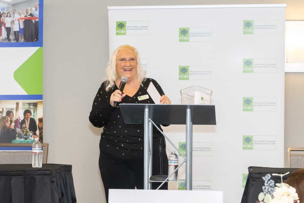 Middle aged woman with blonde hair wearing black, standing at a podium speaking to the audience at a PCBA business panel event