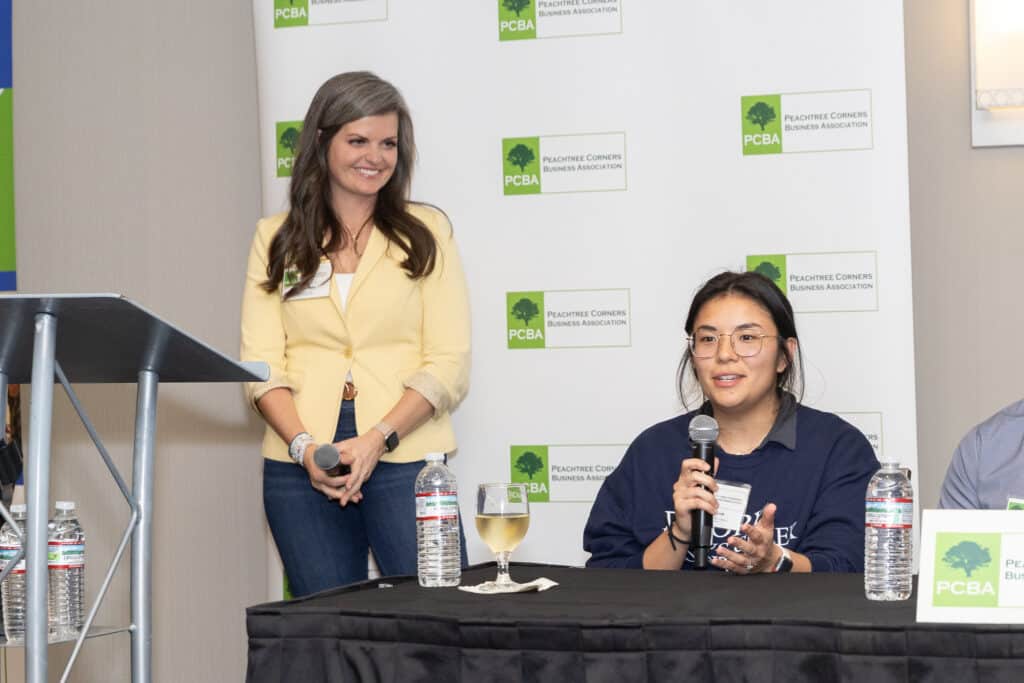 A woman in yellow blouse standing on stage at a business panel event. She's looking at a woman sitting at the panel table next to her and smiling.