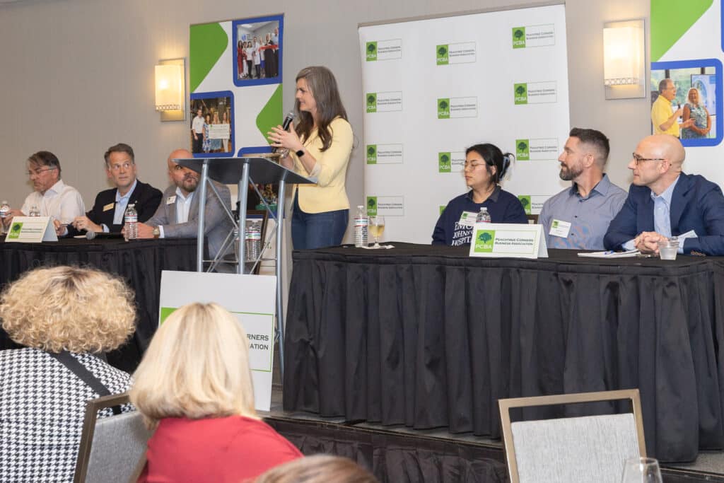 A female host and panel members at a podium and seated at tables in front of an audience at a PCBA panel event on health and wellness.