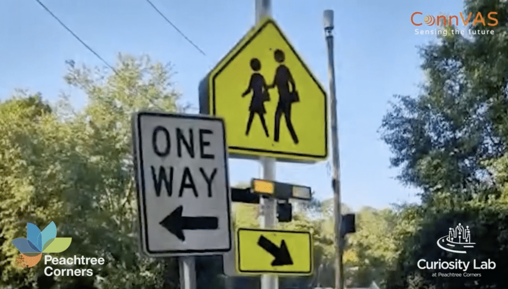 One Way and Pedestrian Crossing signs in a suburban neighborhood with power lines overhead and trees in the background. Logos from city of Peachtree Corners, ConnVAS and Curiosity Lab are on top of the image