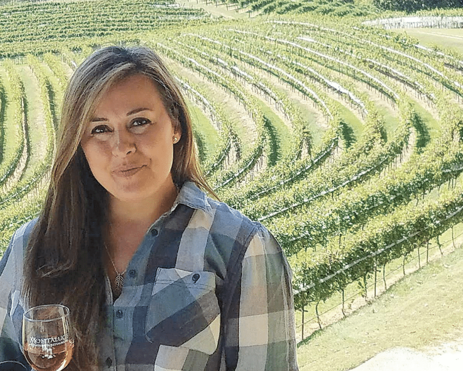 A white woman with blonde hair wearing a blue, white and grey blouse. She's standing outdoors at a vineyard holding a glass of wine.