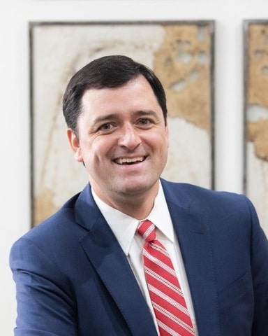 Georgia State Rep. Scott Hilton wearing a blue suit, white button-up shirt and red & white tie. He's smiling and there are framed maps behind him.