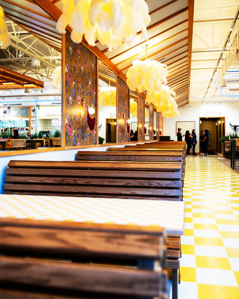 A long view of the dining hall at The Forum Peachtree Corners with dark wood booths, mirrored walls, yellow and white floor and hanging pendant lights. A group of staff members wearing black stand in the background.