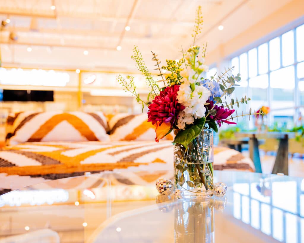 A vase of flowers on a table in front of an orange and white sofa in a well-lit space with lots of windows.