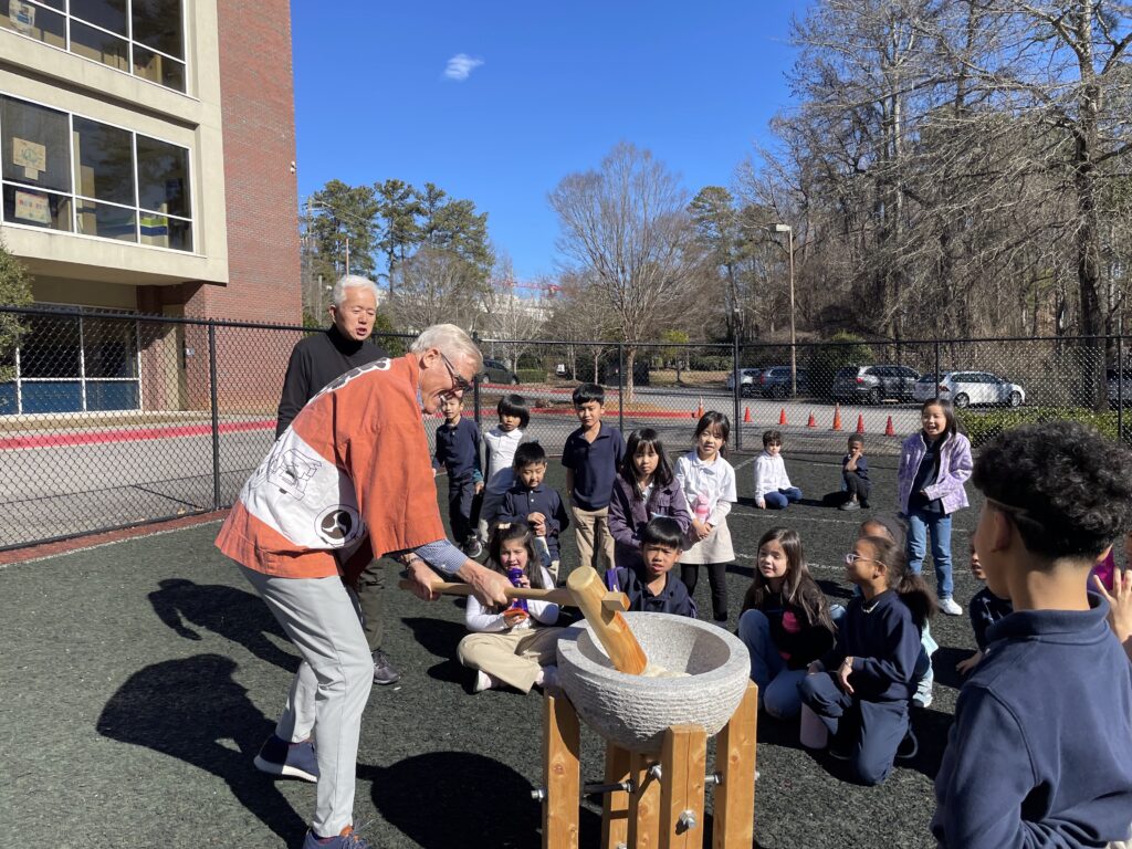 Man and students at a dual-language school pounding rice with a wooden mallet at Japanese new year event.