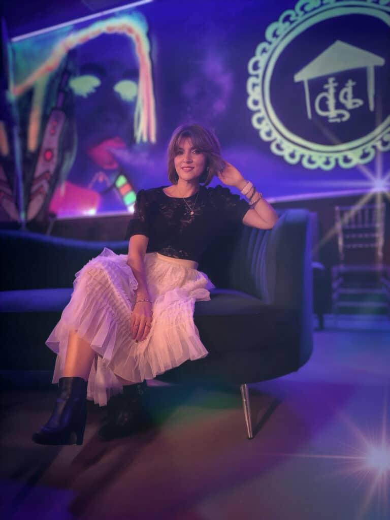 Young woman sitting in a chair in a room with purple walls and lighting. She's wearing a white skirt and black top and looking at the camera.